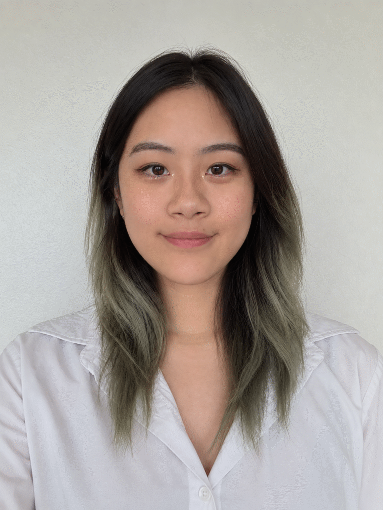 A front-facing portrait of an east asian female presenting human with dark hair fading into muted green-gray ends, wearing a white collared shirt. They're smiling softly at the camera against a plain light-colored background, with subtle makeup, winged eyeliner, and small earrings.