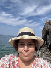 A smiling woman in a straw hat with a black band and a flowered shirt. The sun, sky, lake, and a rock cliff in the background.