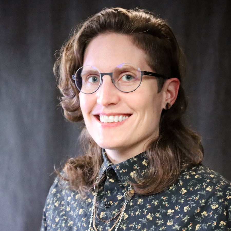 Rua Williams is a white queer presenting person smiling towards the camera in semi profile. They are wearing a floral shirt in black and gold and have round black glasses.