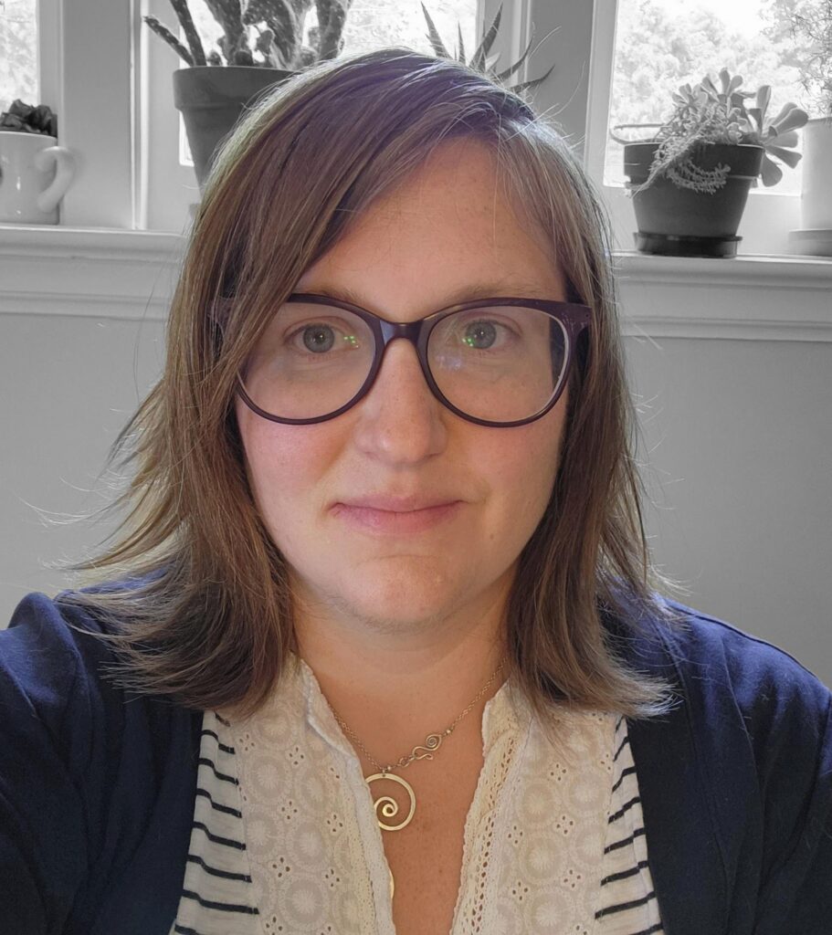 Dani, a white female with shoulder-length reddish brown hair, sits facing the camera with a slight smile. She is wearing large blue rimmed glasses, a black and white striped shirt, and a black blazer. The background is in black and white and shows a wall of windows with the sills covered in various succulents.